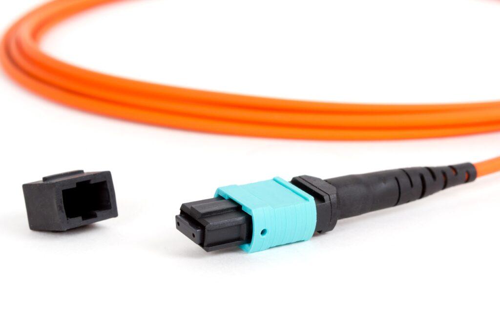 Ein Bild eines MPO-Glasfaserkabels in Orange, verbunden mit einem türkisfarbenen MPO-Stecker, neben einem losen schwarzen Steckergehäuse, auf weißem Hintergrund